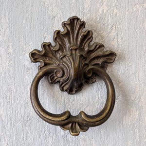 May include: A bronze-toned metal door knocker featuring an ornate, leaf-like backplate with intricate scrollwork details and a circular, curved ring handle that hangs from the center, showcasing a weathered, metallic finish throughout the piece.