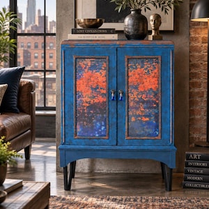 May include: A rectangular blue cabinet with two doors featuring a distressed orange and blue textured finish. The cabinet has a flat top, a base with an arched cutout, and four black tapered legs. Two small metal knobs are centered on the doors.