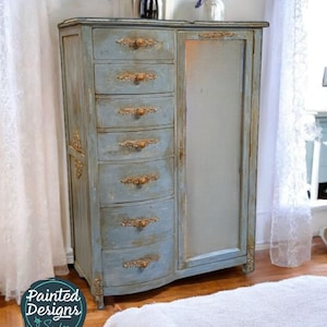 May include: A distressed blue painted armoire with gold accents and a mirrored door. The armoire has eight drawers with ornate gold handles. The armoire is standing on a wooden floor.
