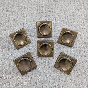 May include: Six antique brass drawer pulls with square bases and circular handles. The pulls are arranged on a light gray carpet. The hardware has a vintage aesthetic.
