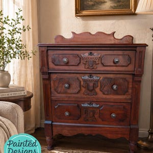May include: A dark brown antique dresser with three drawers and ornate carvings. The dresser has a decorative top and round drawer pulls. The image also includes a painting, a lamp, and a plant in a vase. The text "Painted Designs by Sondra" is in the lower left corner.