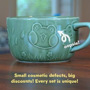 May include: A sage green ceramic mug featuring a frog face design. The mug has a curved handle and is decorated with raindrops and flowers. Text at the bottom reads "Small cosmetic defects, big discounts! Every set is unique!"