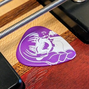 Pu&ograve; includere: Un plettro per chitarra viola con un'illustrazione bianca di un personaggio dei cartoni animati che fa surf su un'onda. Il plettro &egrave; appoggiato su una superficie di legno, con una corda argentata e hardware nero.