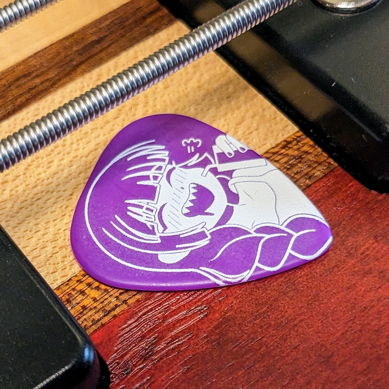 Pu&ograve; includere: Un plettro per chitarra viola con un'illustrazione bianca di un personaggio dei cartoni animati che fa surf su un'onda. Il plettro &egrave; appoggiato su una superficie di legno, con una corda argentata e hardware nero.