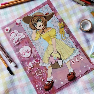 May include: A pink plastic folder featuring a colorful anime illustration of a girl in a yellow dress and a brown hat. The folder has decorative stickers and Japanese text. The girl is holding a basket with a teddy bear.