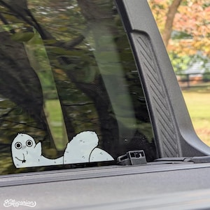Puede incluir: Una pegatina de vinilo blanco con una criatura de dibujos animados en forma de gusano con ojos grandes y una pequeña sonrisa. La pegatina está aplicada en la ventana de un coche, con el texto "Kittygarian".
