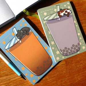 May include: Two small blue and green notebooks with a cat sleeping on top of a boba tea. The notebooks have a star pattern and the boba tea has brown and white boba pearls.