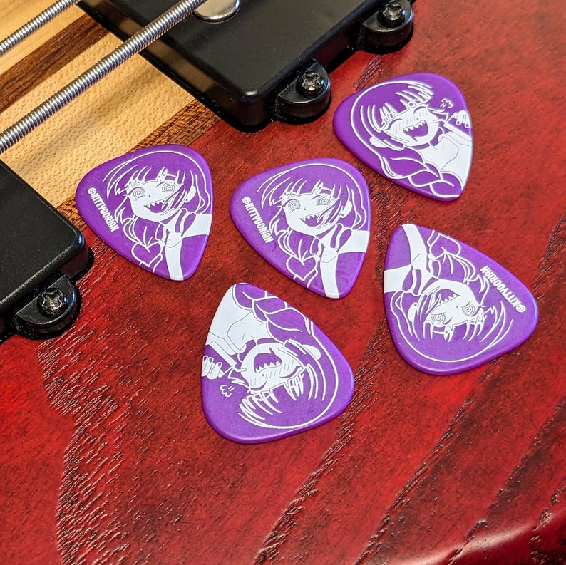 Pu&ograve; includere: Sei plettri per chitarra viola con un'illustrazione bianca in stile anime di un personaggio con un cuore e il testo "@KITTYOORIAN". I plettri sono disposti su una superficie di legno, vicino a un basso.