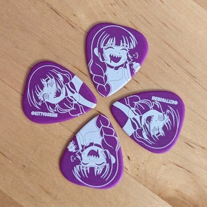 Pu&ograve; includere: Quattro plettri per chitarra viola con un'illustrazione bianca di una ragazza con trecce e un'espressione giocosa. Il testo "@KITTYGORIAN" &egrave; stampato su ogni plettro. I plettri sono disposti su una superficie di legno.