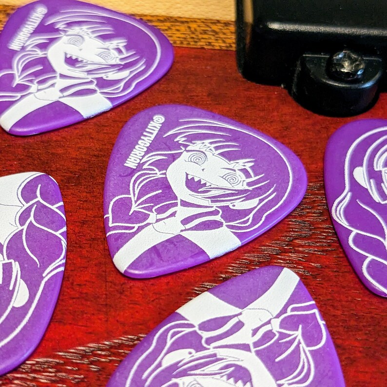 Pu&ograve; includere: Plettri per chitarra viola con un design di personaggio dei cartoni animati bianco e la scritta "KITTYGORIAN". I plettri sono sparsi su una superficie rossa, con un oggetto nero sullo sfondo. I plettri hanno una forma triangolare.