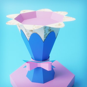 Puede incluir: Un recipiente de papel con forma de flor con base azul e interior rosa. El recipiente tiene un lazo rosa y se asienta sobre una base geométrica rosa y azul. El borde superior tiene un estampado floral.