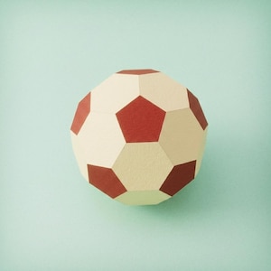 Puede incluir: Una pelota geométrica construida en papel, con un patrón repetitivo de hexágonos color crema y pentágonos de color rojo oscuro. El objeto tiene una textura mate y una superficie estructurada de múltiples facetas, creando una forma esférica precisa.