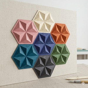 Pode incluir: Uma peça de arte de parede com sete painéis hexagonais dispostos em grupo sobre um fundo de tecido bege claro. Cada hexágono apresenta um padrão geométrico de estrela tridimensional em cores distintas: creme, rosa, laranja, azul, azul-petróleo, verde e cinza.