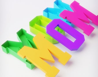 Puede incluir: La palabra "MAMÁ" escrita en letras 3D de colores. Las letras están hechas de papel y están apiladas una encima de la otra. Las letras son verdes, amarillas, moradas, rosas y azules.