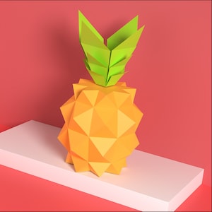 May include: A papercraft pineapple with a yellow body and green leaves. The pineapple is sitting on a white shelf against a pink background.