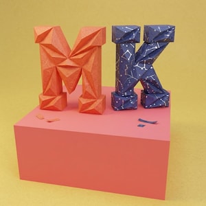 Puede incluir: Una letra M y una letra K tridimensionales hechas de papel plegado. La letra M es de color naranja sólido con una superficie geométrica facetada. La letra K es de color azul oscuro con un patrón de constelaciones blanco y textura facetada.