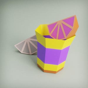 Op de afbeelding: Een geometrisch papieren cocktailglas met gele, paarse en oranje secties. Een papieren schijfje citrusvrucht ligt bovenop het glas, met een ander schijfje ernaast. De achtergrond is lichtgroen.