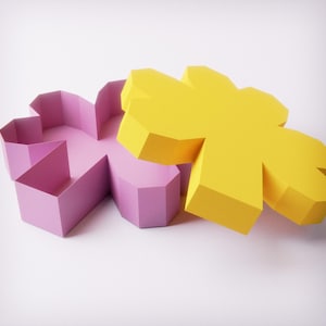 Peut inclure: Deux boîtes géométriques en forme de fleur. L'une est jaune vif, l'autre rose tendre. Les boîtes ont un design à facettes, avec de multiples côtés créant les pétales de la fleur. Le fond est une surface blanche et propre.