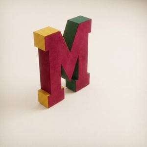 May include: A three-dimensional letter M standing upright. The front face is a deep magenta color, the side edges are a dark forest green, and the top and bottom edges are a bright mustard yellow. The letter has a blocky, geometric structure.