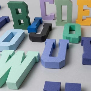 Può includere: Una collezione di lettere tridimensionali a blocchi in vari colori, tra cui verde, blu, giallo, nero e lavanda. Ogni lettera presenta una struttura geometrica spessa con bordi netti e una finitura opaca, disposte in modo sparso.