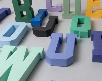 Puede incluir: Una colección de letras en bloque tridimensionales de varios colores, incluyendo verde, azul, amarillo, negro y morado. Estos caracteres alfabéticos gruesos con acabado mate están dispuestos de forma dispersa, mostrando formas geométricas.