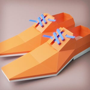 May include: A pair of orange paper shoes with blue shoelaces. The shoes have a geometric design and white soles. The shoes are displayed on a light brown surface.