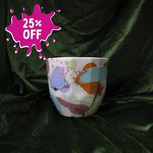 May include: A white ceramic planter with a faceted surface featuring abstract painted shapes in lavender, light blue, orange, and pink, accented with small gold-colored speckles. A pink graphic in the upper left corner displays the text 25% OFF.