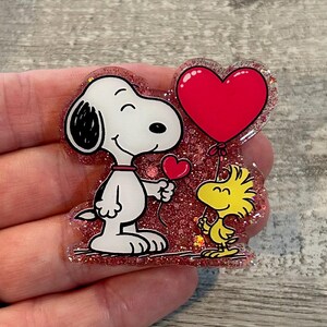 May include: A Snoopy and Woodstock-themed pin. Snoopy, in black and white, holds a small red heart. Woodstock, in yellow, holds a large red heart balloon. The pin has a glittery red background and is held by a hand.