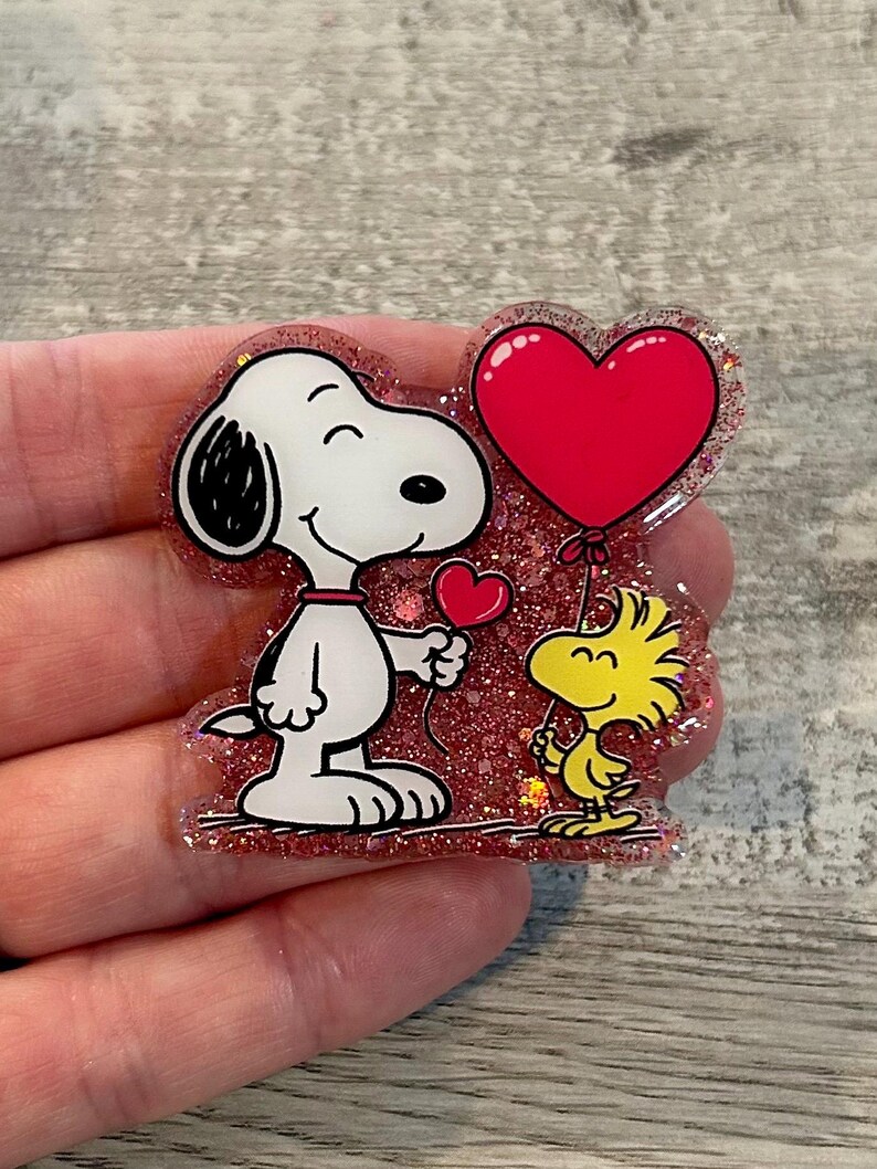 May include: A Snoopy and Woodstock-themed pin. Snoopy, in black and white, holds a small red heart. Woodstock, in yellow, holds a large red heart balloon. The pin has a glittery red background and is held by a hand.