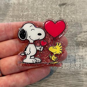 May include: A glittery, heart-shaped pin featuring Snoopy and Woodstock. Snoopy, in black and white, holds a small red heart, while Woodstock holds a large red heart balloon. The pin has a pink glitter border.