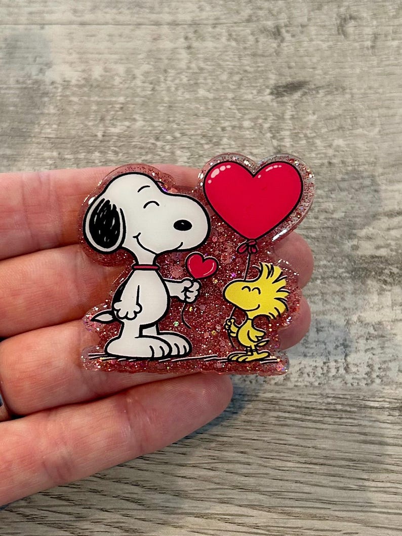 May include: A glittery, heart-shaped pin featuring Snoopy and Woodstock. Snoopy, in black and white, holds a small red heart, while Woodstock holds a large red heart balloon. The pin has a pink glitter border.