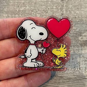 May include: A glittery, heart-shaped decoration featuring Snoopy and Woodstock. Snoopy, in black and white, holds a small heart, while Woodstock holds a large red heart balloon. The edges are outlined in red glitter.