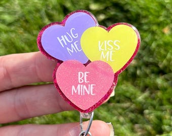 May include: A badge reel featuring three heart-shaped charms with glitter edges. The charms are purple, yellow, and pink, with the phrases "HUG ME", "KISS ME", and "BE MINE" in white lettering. The reel has a clear plastic clip.