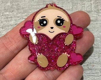 May include: A glittery pink sloth-shaped charm holding a heart. The sloth has a tan face, large black eyes, and pink cheeks. The heart is also pink and glittery, with smaller pink hearts around the edges. The charm is a cute, whimsical design.
