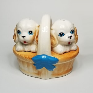 May include: A ceramic salt and pepper shaker set shaped like two puppies in a brown wicker basket with a blue bow.