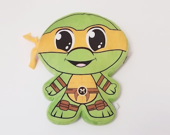 May include: Green and orange plush toy of a Teenage Mutant Ninja Turtle with a big smile and large eyes. The turtle is wearing a brown belt with a letter 'M' on it.