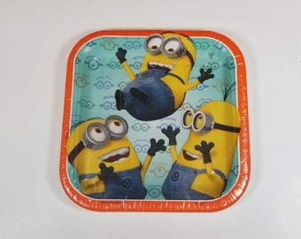 May include: Square paper plate featuring three Minion characters in a playful design. The plate has a teal background with a pattern of Minion goggles and an orange border. The Minions are yellow with blue overalls and goggles.