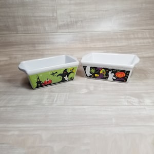May include: Two white ceramic loaf pans with Halloween themed designs. One pan has a green background with a ghost, a cat, and a pumpkin. The other pan has a black background with a witch, a ghost, and a pumpkin.