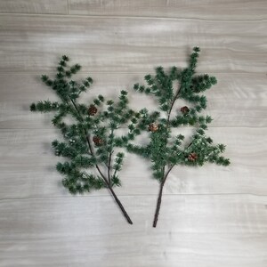 May include: Two sprigs of artificial pine branches with brown pine cones. The branches are green and have a frosted look.
