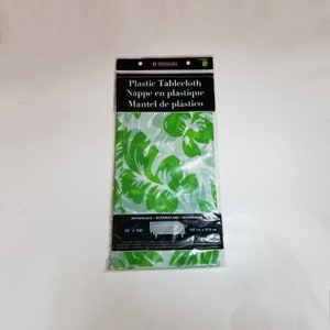 May include: A green plastic tablecloth with a tropical leaf pattern. The tablecloth is rectangular and measures 54 inches by 108 inches. The packaging also includes the text "Plastic Tablecloth", "Nappe en plastique", and "Mantel de plástico".