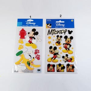 May include: Two packages of Disney Mickey Mouse stickers. The first package has a red fire hydrant, a yellow dog, and Mickey Mouse. The second package has Mickey Mouse in different poses and a few stars.