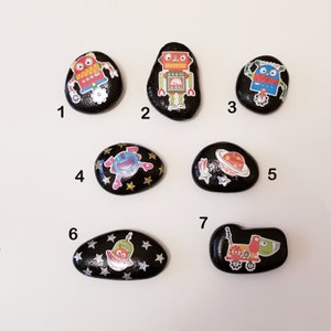 May include: Seven painted rocks with different space-themed designs. The rocks are black with gold glitter and feature images of robots, planets, and a spaceship.