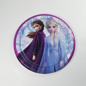 May include: A round paper plate with a purple border features a colorful illustration of two girls from the animated movie Frozen. The girls are wearing blue and purple dresses and have long blonde and brown hair. The background is a light blue with white floral designs.