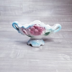 May include: A white ceramic bowl with a blue and pink floral design. The bowl has two handles and sits on a pedestal.