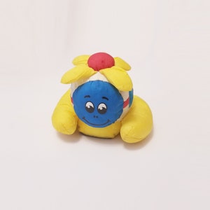 May include: A yellow plush toy shaped like a flower with a blue face and a red and white center. The toy has a smiling face with two large eyes.