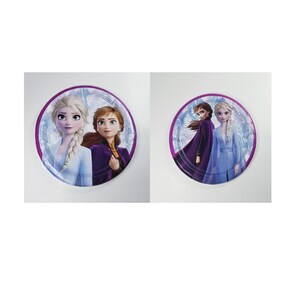 May include: Two round paper plates featuring images of Elsa and Anna from the movie Frozen. The plates have a purple border and a blue and white snowflake pattern.