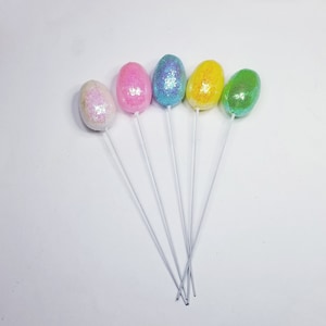 May include: Five pastel coloured Easter eggs on white sticks. The eggs are pink, blue, yellow, and green.