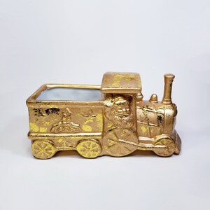 May include: Gold ceramic train planter with a Santa Claus figure on the front. The planter has a white interior and is decorated with stars and holly.