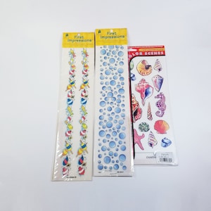 May include: Three packs of stickers featuring a beach theme. The first pack has a blue and white border with a repeating pattern of beach items like ice cream cones, seashells, and fish. The second pack has a blue and white border with a repeating pattern of blue bubbles. The third pack has a red and white border with a variety of sea life stickers, including seashells, starfish, and a seahorse. The package says "Color Scenes Ice Cream Stick 23 Sea Shells C501NC"