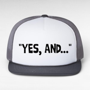 Peut inclure: Casquette de camionneur avec un panneau avant blanc et un dos en filet gris. La casquette affiche la phrase "YES, AND..." en lettres noires grasses.